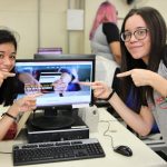 Foto de duas jovens apontando para a tela de um computador e sorrindo