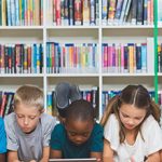 Foto de dois meninos e uma menina deitados no chão, mexendo em tablets. Ao fundo, há uma estante com diversos livros