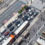 Foto aérea de uma avenida com carros, motos e ônibus parados. Há pedestres atravessando a faixa de segurança e outras pessoas nas calçadas e em um ponto de ônibus.